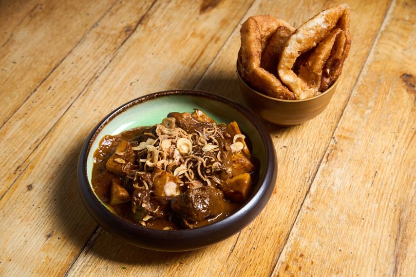 Rich brown curry with fried shallots in a green bowl, with a side of crispy flatbread.
