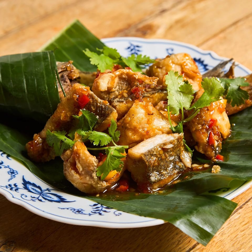Crispy fried fish pieces with sweet chili sauce and cilantro, served on a banana leaf.