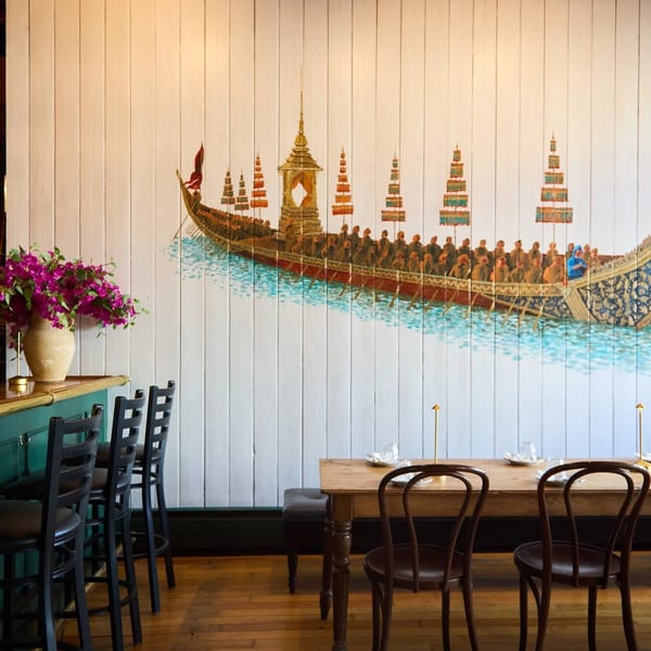 Restaurant interior with a long wooden table, bar stools, and a large mural of a ceremonial boat.