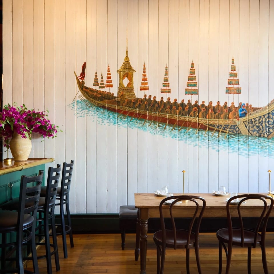 Restaurant interior with a long wooden table, bar stools, and a large mural of a ceremonial boat.