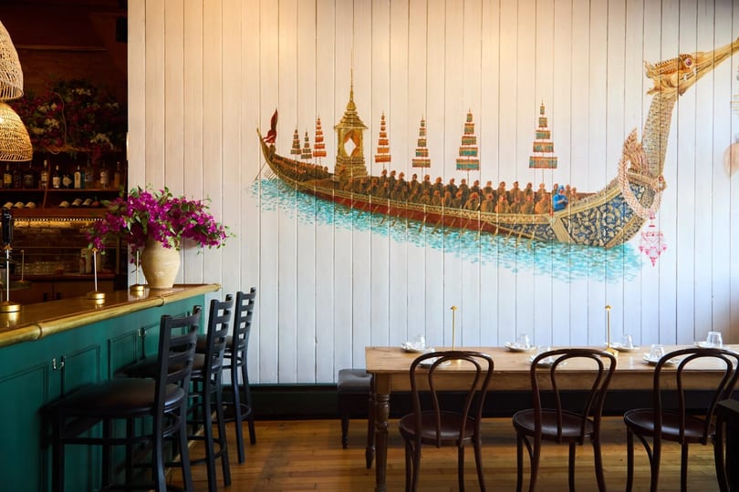 Restaurant interior with a long wooden table, bar stools, and a large mural of a ceremonial boat.