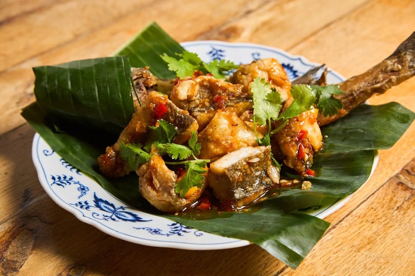 Crispy fried fish pieces with sweet chili sauce and cilantro on a banana leaf-lined plate.