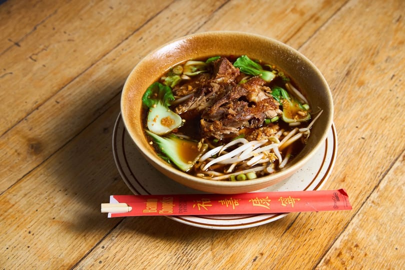A bowl of ramen with pork, bok choy, and bean sprouts on a wooden table with chopsticks.