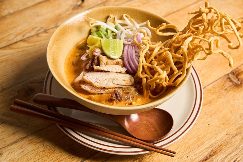 A bowl of Khao Soi with chicken, crispy noodles, red onion, lime, and bean sprouts.