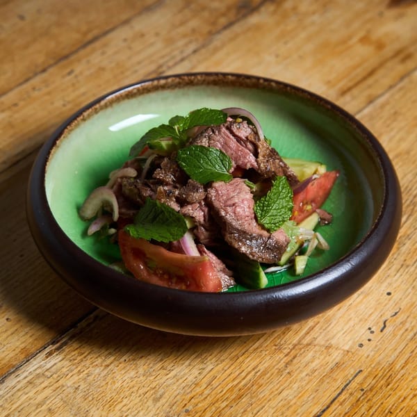 Thai beef salad with mint, red onion, tomatoes, and cucumbers in a green bowl on a wooden table.
