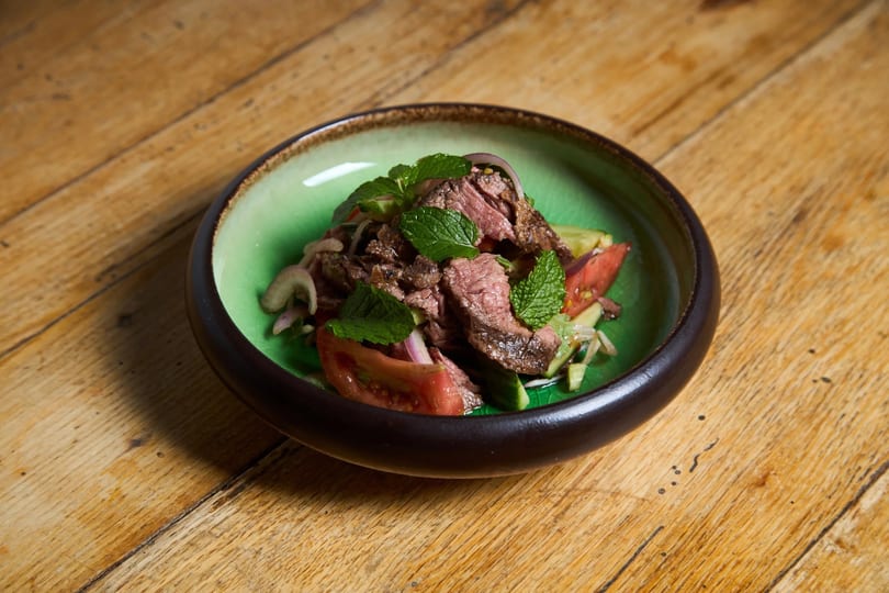 Thai beef salad with mint, red onion, tomatoes, and cucumbers in a green bowl on a wooden table.