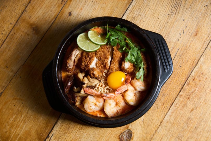 A black bowl of ramen with crispy chicken, shrimp, egg yolk, lime, and cilantro on wood.