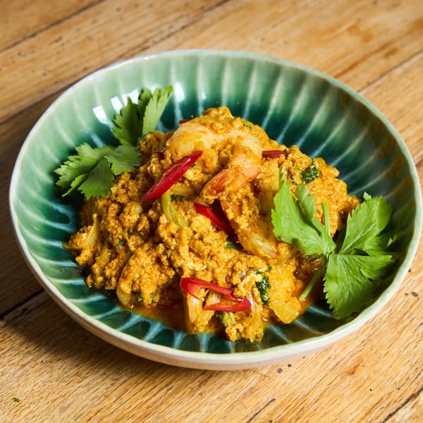 A green bowl of yellow curry with shrimp, red chili slices, and cilantro on a wooden table.