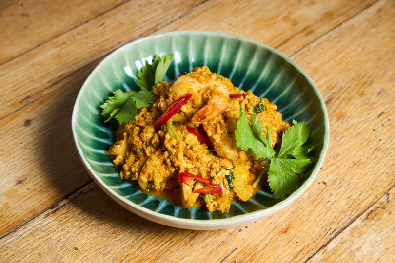 A green bowl of yellow curry with shrimp, red chili slices, and cilantro on a wooden table.