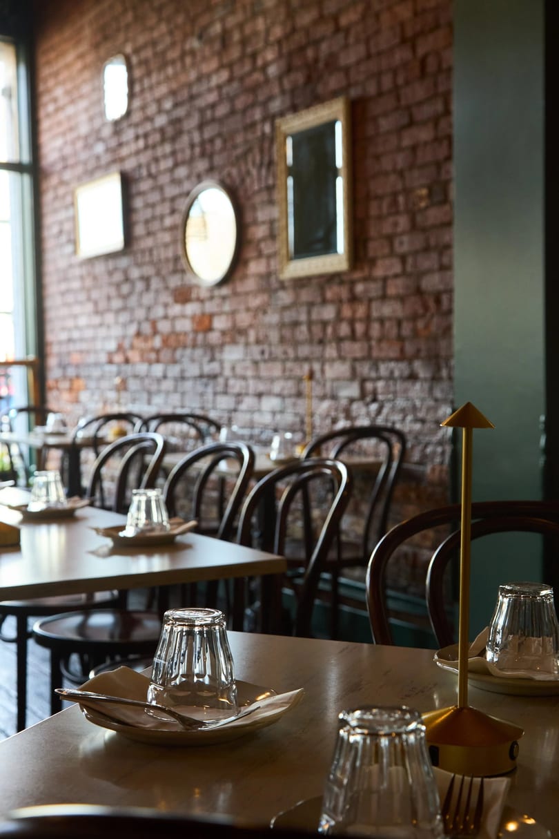 Restaurant interior with set tables, dark wooden chairs, and a brick wall adorned with mirrors.